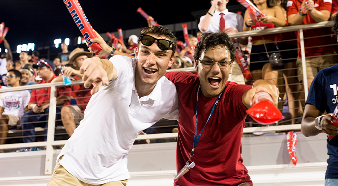 Two male students during an FAU football game smiling and pointing at the camera